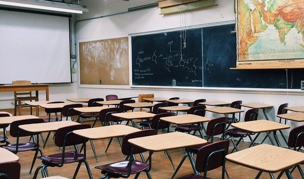 Klassenraum mit vielen SchulTischen und Stühlen. Seitlich Hängt eine große Tafel und ein Teil einer Weltkarte.