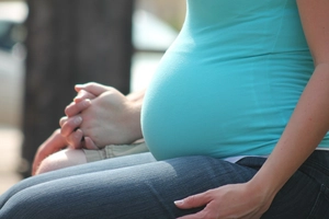 Im Vordergrund sitzt eine schwangere Frau. Nur ihr Bauch und ihre Beine sind zu sehen. Im Hintergrund sitzt noch eine weitere Person.
