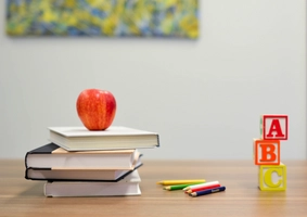 Mehrer Bücher liegen aufeinander auf dem Tisch. Auf den Büchern liegt ein Apfel. Neben den Büchern liegen Buntstifte. Neben den Buntstiften sind 3 Baukötzer mit den Buchstaben A, B und C aufeinander gestapelt.