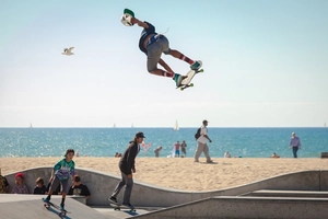 An einem Skaterpark an einem Strand fahren 3 Jugendliche Skateboard. Einer von Ihnen macht gerade einen Stunt in der Luft.
